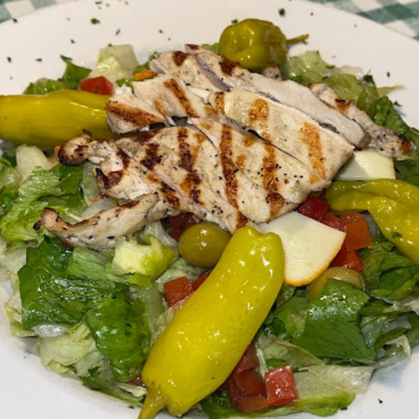 Grilled chicken salad with lettuce, tomatoes, olives, and yellow peppers on a white plate.
